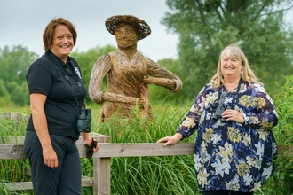 Two women stood smiling to camera in front of a reed bed with a giant willow weaved man in the background