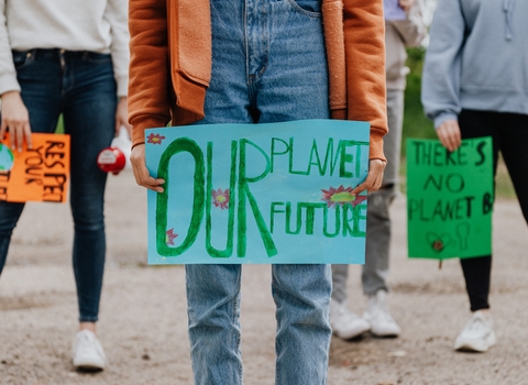 An image of a young woman wearing jeans, trainers and a orange jumper holding a sign that reads 'Our planet, our future!'