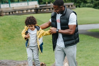 A man and his son are playing outdoors, the son is walking on a  log whilst the father holds his hand. They are both smiling