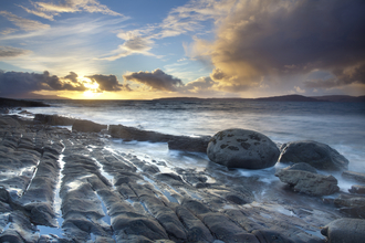 A coastal landscape, with the sea gently lapping at smooth rocks as the sun sets behind scattered clouds