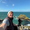 Joe is smiling in a selfie on a rocky coastal cliff, with a spotting scope on a tripod beside them, overlooking a blue sea and small rocky islands under a clear sky.
