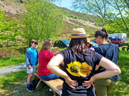 A woman wearing a Stand for Nature Wales branded tshirt with her back to the camera. There are children and adults doing crafts in the background.