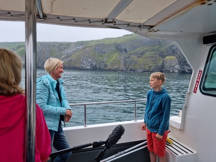 Young boy talks to a woman whilst standing on a boat with cliffs and the ocean in the background