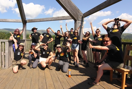 A photo of 15 people all wearing black tshirts with Stand for Nature Wales branding doing silly poses in a bird observatory