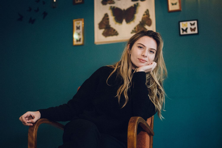 Sian Eleri sat on a wooden chair wearing a black jumper in front of a green wall with photos of butterflies 
