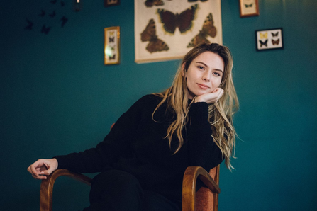 Sian Eleri sat on a wooden chair wearing a black jumper in front of a green wall with photos of butterflies