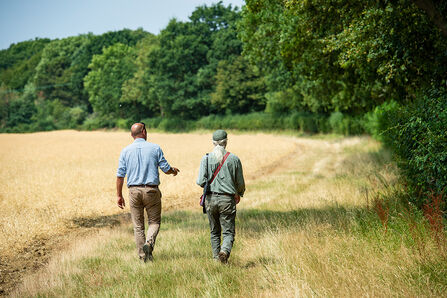 Two men, a farm advisor and a farmer, are walking down the edge of a field, alongside a line of hedges and tall trees