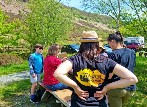 A woman wearing a Stand for Nature Wales branded tshirt with her back to the camera. There are children and adults doing crafts in the background.