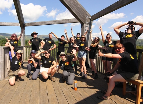 A photo of 15 people all wearing black tshirts with Stand for Nature Wales branding doing silly poses in a bird observatory